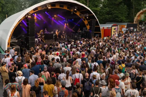 Ein Foto von einer Festivalb&uuml;hne mit vielen Menschen, die davor stehen. Die Festivalb&uuml;hne ist violett beleuchtet und &uuml;berdacht.